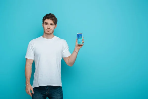 Jeune homme mécontent montrant smartphone avec application de magasinage en ligne à l'écran sur fond bleu — Photo de stock