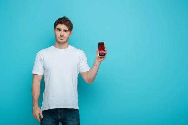 Joven disgustado mostrando teléfono inteligente con cursos de comercio en pantalla sobre fondo azul - foto de stock