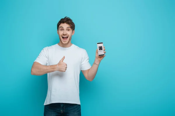 KYIV, UKRAINE - APRIL 23, 2019: excited young man showing thumb up while holding smartphone with Uber app on screen on blue background — Stock Photo