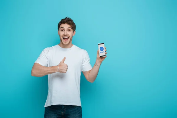 KYIV, UKRAINE - APRIL 23, 2019: excited young man showing thumb up while holding smartphone with Zoom app on screen on blue background — Stock Photo