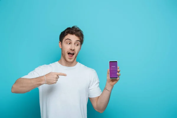 KYIV, UKRAINE - APRIL 23, 2019: excited guy pointing at smartphone with Instagram app on screen on blue background — Stock Photo