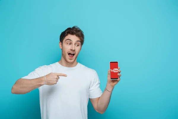 KYIV, UKRAINE - APRIL 23, 2019: excited guy pointing at smartphone with Youtube app on screen on blue background — Stock Photo