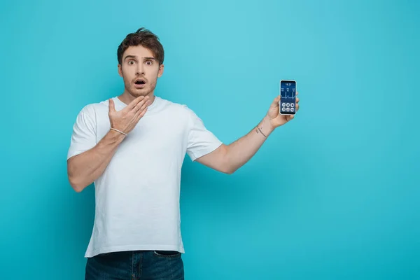 Chocado chico sosteniendo la mano cerca de la cara mientras muestra el teléfono inteligente con la aplicación de ritmo cardíaco sobre fondo azul - foto de stock