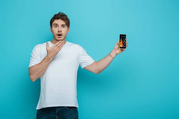 Cara chocado segurando a mão perto do rosto ao mostrar smartphone com aplicativo de análise de negócios no fundo azul — Fotografia de Stock