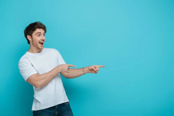 Cheerful young man pointing with fingers while looking away on blue background — Stock Photo