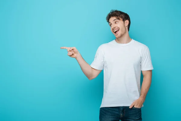 Sorrindo cara segurando mão no bolso, olhando para longe e apontando com o dedo no fundo azul — Fotografia de Stock