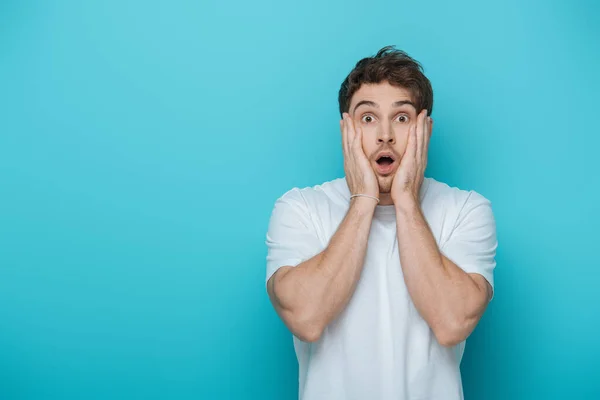 Shocked man touching face while looking at camera on blue background — Stock Photo