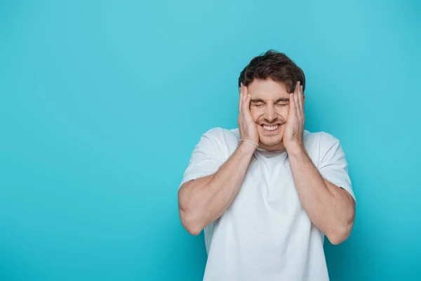 Verängstigter junger Mann berührt Gesicht, während Grimassen mit geschlossenen Augen auf blauem Hintergrund — Stockfoto