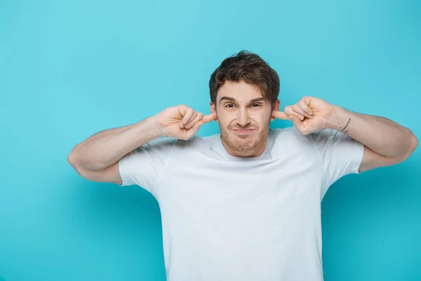 Displeased young man plugging ears with fingers while looking at camera on blue background — Stock Photo