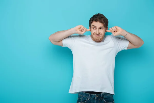 Joven disgustado tapando las orejas con los dedos mientras mira hacia otro lado sobre fondo azul - foto de stock