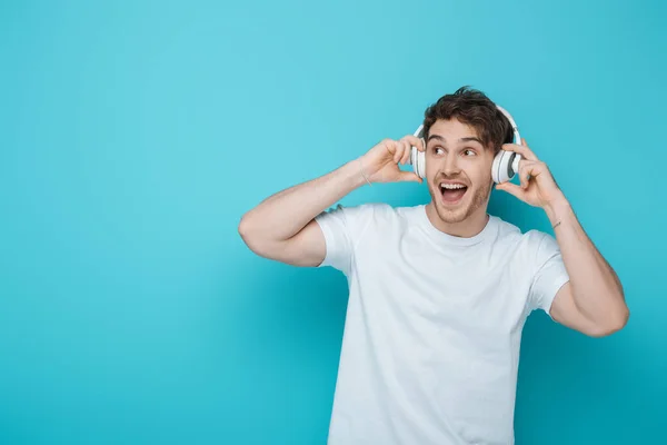 Chico emocionado tocando auriculares inalámbricos y mirando hacia otro lado sobre fondo azul - foto de stock