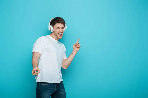 Збуджений молодий чоловік танцює і співає в бездротових навушниках на синьому фоні — стокове фото