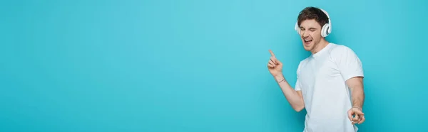 Panoramic crop of excited young man dancing and singing in wireless headphones on blue background — Stock Photo