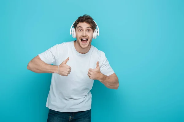 Excited young man in wireless headphones showing thumbs up while looking at camera on blue background — Stock Photo
