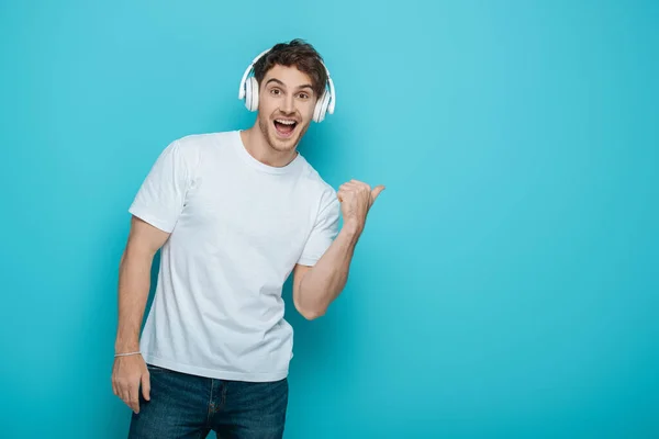 Excited young man in wireless heaphones pointing with thumb while looking at camera on blue background — Stock Photo