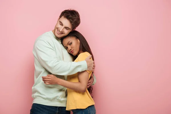 Linda menina americana africana olhando para a câmera enquanto namorado feliz abraçando-a no fundo rosa — Fotografia de Stock