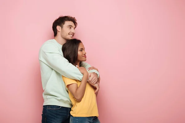Feliz chico abrazando sonriente novia mientras mira hacia otro lado juntos en rosa fondo - foto de stock