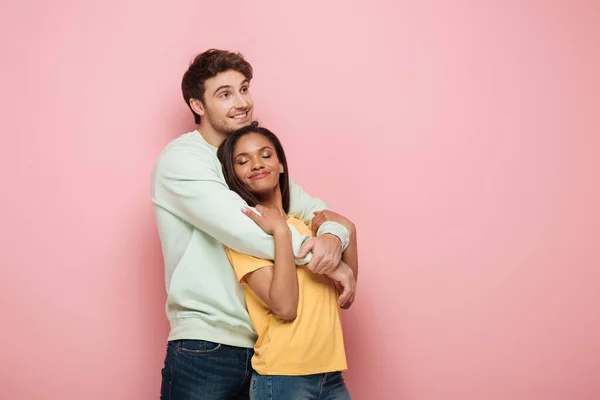 Feliz chico abrazando sonriente novia mientras mira hacia otro lado en rosa fondo - foto de stock