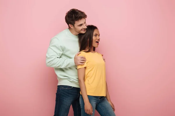 Smiling man touching shoulders of amazed girlfriend looking away on pink background — Stock Photo