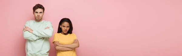 Horizontal image of offended interracial couple standing with crossed arms while looking at camera on pink background — Stock Photo