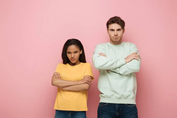 Pareja interracial disgustado de pie con los brazos cruzados mientras mira a la cámara en el fondo rosa - foto de stock