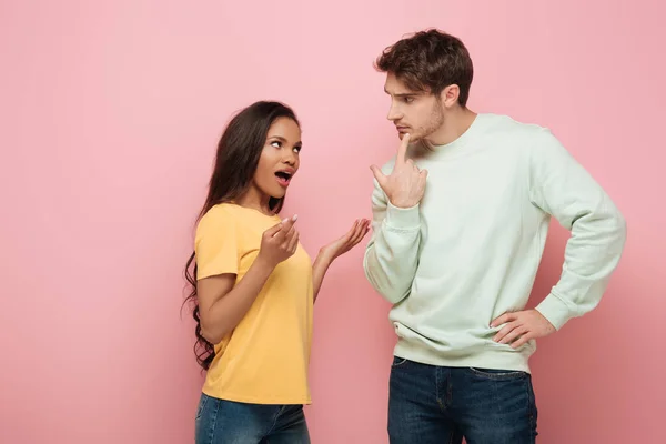 Serious guy touching chin while looking at african american girlfriend quarreling on pink background — Stock Photo