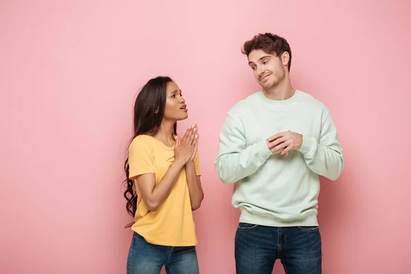 Bella ragazza afroamericana con le mani in preghiera guardando sorridente, fidanzato scettico su sfondo rosa — Foto stock