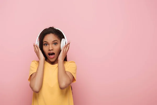 Chocado afro-americano menina tocando sem fio fones de ouvido enquanto olha para a câmera no fundo rosa — Fotografia de Stock