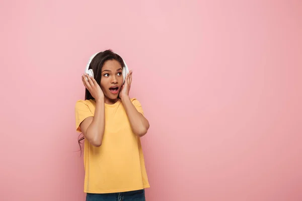 Scioccato afro-americano ragazza toccando cuffie wireless mentre guardando altrove su sfondo rosa — Foto stock