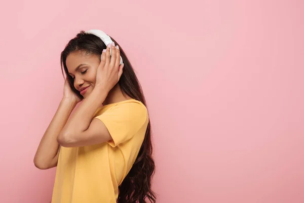 Chica afroamericana sonriente escuchando música en auriculares inalámbricos con los ojos cerrados aislados en rosa - foto de stock