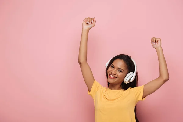 Glückliches afrikanisches amerikanisches Mädchen in drahtlosen Kopfhörern, das mit erhobenen Händen tanzt, während es in die Kamera auf rosa Hintergrund schaut — Stockfoto