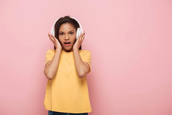 Shocked african american girl touching wireless headphones while looking at camera on pink background — Stock Photo