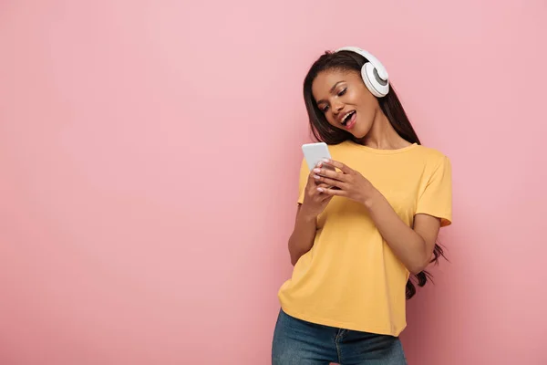 Chica afroamericana feliz en auriculares inalámbricos charlando en el teléfono inteligente sobre fondo rosa - foto de stock