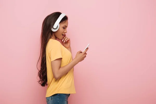 Vista lateral de la niña afroamericana reflexiva en auriculares inalámbricos utilizando teléfono inteligente aislado en rosa - foto de stock