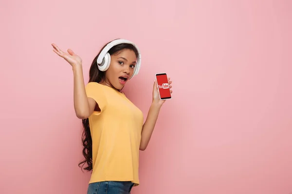 KYIV, UKRAINE - 23 AVRIL 2019 : fille afro-américaine excitée dans des écouteurs sans fil montrant smartphone avec application Youtube sur fond rose — Photo de stock