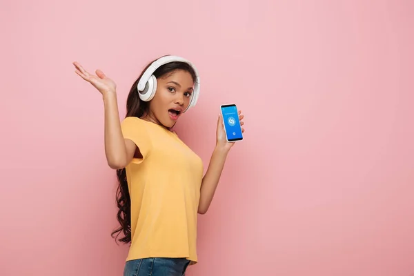 KYIV, UCRANIA - 23 DE ABRIL DE 2019: chica afroamericana emocionada con auriculares inalámbricos que muestran el teléfono inteligente con la aplicación Shazam sobre fondo rosa - foto de stock
