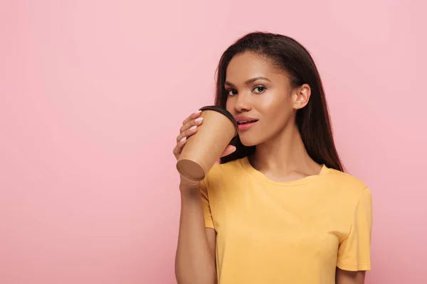 Atraente menina afro-americana beber café para ir enquanto olha para a câmera isolada em rosa — Fotografia de Stock