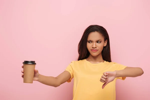 Unzufriedene afrikanisch-amerikanische Mädchen zeigt Daumen nach unten, während sie Kaffee hält, um auf rosa Hintergrund zu gehen — Stockfoto