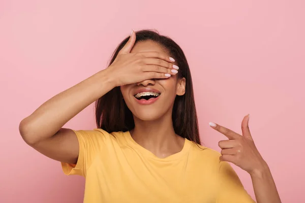 Жизнерадостная африканская американка, закрывающая глаза рукой, указывая пальцем на брекеты на зубах, изолированные розовым. — стоковое фото