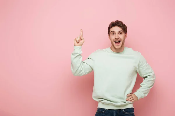 Возбужденный парень, показывающий знак идеи, стоя с рукой на бедре и глядя в камеру на розовом фоне — стоковое фото