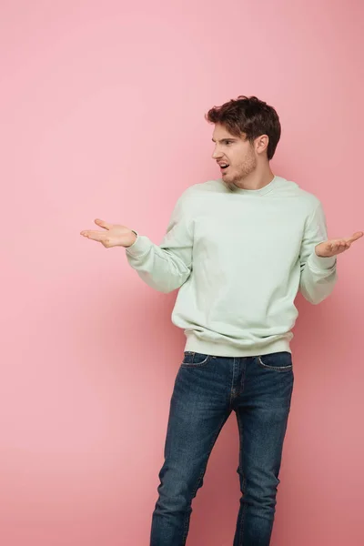 Displeased young man standing with open arms while looking away on pink background — Stock Photo