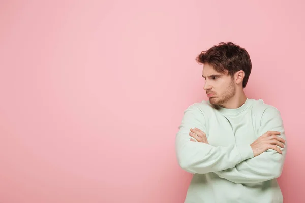 Ofendido joven mirando hacia otro lado mientras estaba de pie con los brazos cruzados sobre fondo rosa - foto de stock