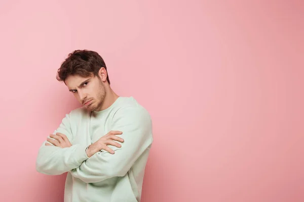 Offended young man looking at camera while standing with crossed arms on pink background — Stock Photo