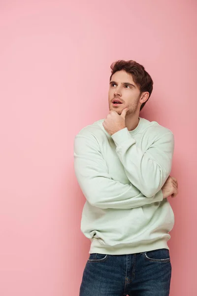 Verträumter junger Mann berührt Kinn beim Wegschauen auf rosa Hintergrund — Stockfoto