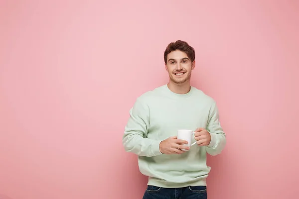 Handsome young man holding white cup while smiling at camera on pink background — Stock Photo