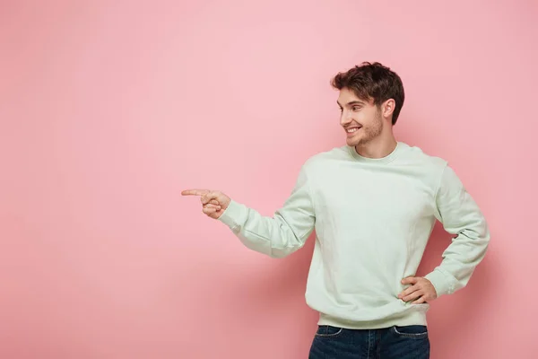 Alegre joven mirando hacia otro lado y señalando con el dedo mientras sostiene la mano en la cadera sobre fondo rosa - foto de stock