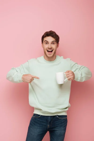 Aufgeregter Typ, der mit dem Finger auf weiße Tasse zeigt, während er in die Kamera auf rosa Hintergrund schaut — Stockfoto