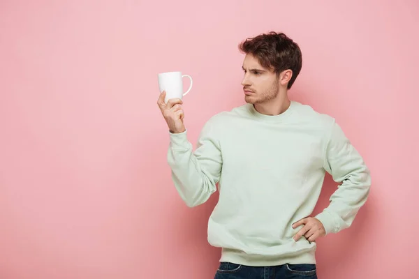 Nachdenklicher junger Mann blickt auf weiße Tasse, während er die Hand auf Hüfte auf rosa Hintergrund hält — Stockfoto
