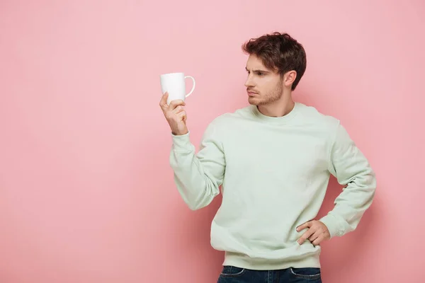 Hombre joven reflexivo mirando taza blanca mientras sostiene la mano en la cadera sobre fondo rosa - foto de stock