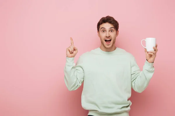 Hombre emocionado mostrando gesto de idea mientras sostiene la taza blanca y mirando a la cámara en el fondo rosa - foto de stock